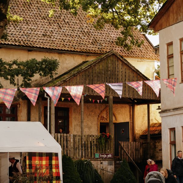 Festīva aina Alsungā ar senām ēkām un cilvēkiem, virs kuras karājas pleda raksta karodziņu virtene. Priekšplānā balta telts, bet pie koka nojumes redzama zīme ar uzrakstu "i ieeja pils".