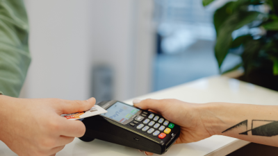 Divas rokas veic maksājumu, ievietojot kredītkarti maksājumu terminālī, ko tur otra roka. Uz termināļa ekrāna redzams uzraksts "PayTel".