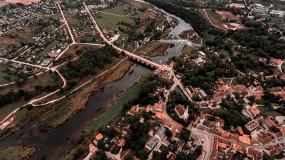Gaisa skats uz Kuldīgas pilsētu ar Ventas upi, veco ķieģeļu tiltu un plašo Ventas rumbu. Abpus upei redzamas ēkas ar sarkanīgiem jumtiem, ko ieskauj zaļi lauki un koki.