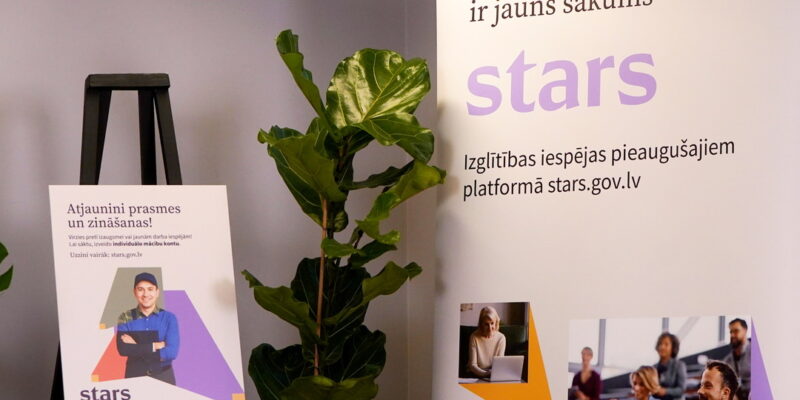 Divi plakāti reklamē izglītības platformu pieaugušajiem "stars.gov.lv". Kreisais plakāts ar uzrakstu "Atjaunini prasmes un zināšanas!" attēlo smaidīgu vīrieti un tekstu "Virzies pretī izaugsmei vai jaunām darba iespējām! Lai sāktu, izveido individuālo mācību kontu. Uzzini vairāk: stars.gov.lv". Labais plakāts, kurā lieli burti veido vārdu "stars", satur uzrakstus "Katrs punkts uz taisnes ir jauns sākums" un "Izglītības iespējas pieaugušajiem platformā stars.gov.lv", kā arī attēlus ar cilvēkiem, kas mācās vai strādā ar datoru. Starp plakātiem stāv liels zaļš telpaugs.