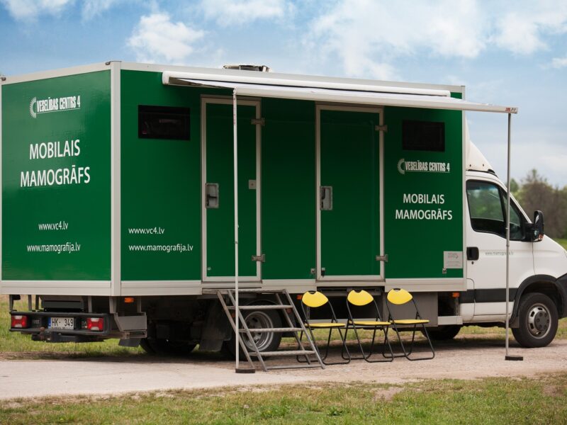 Zaļi-balts kravas auto ar numura zīmi HK-349, uz kura sāniem rakstīts "VESELĪBAS CENTRS 4" un "MOBILAIS MAMOGRĀFS", kā arī "www.vc4.lv" un "www.mamografija.lv", stāv ārā ar izvilktu nojumi, kāpnēm un trim dzelteniem saliekamiem krēsliem.