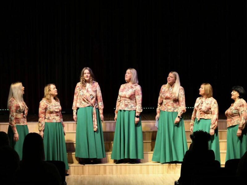 Septiņas sievietes saskaņotos tērpos – ziedu raksta blūzēs un garos, zaļos svārkos – uzstājas uz skatuves. Viņas stāv rindā pret tumšu fonu, dažas skatās viena uz otru, dažas uz priekšu, un viena sieviete dzied ar atvērtu muti. Priekšplānā redzami skatītāju silueti.