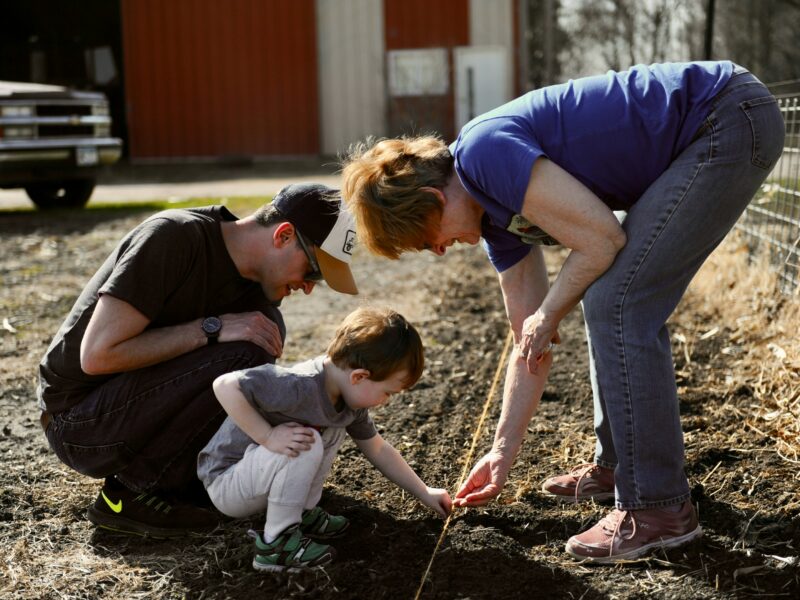 Trīs cilvēki – vīrietis, mazs bērns un sieviete – tupus stāda sēklas augsnē, izmantojot auklu kā vadlīniju. Sieviete rāda bērnam, kā to darīt. Saules apgaismotā fonā redzams sarkans šķūnis un kravas automašīna.